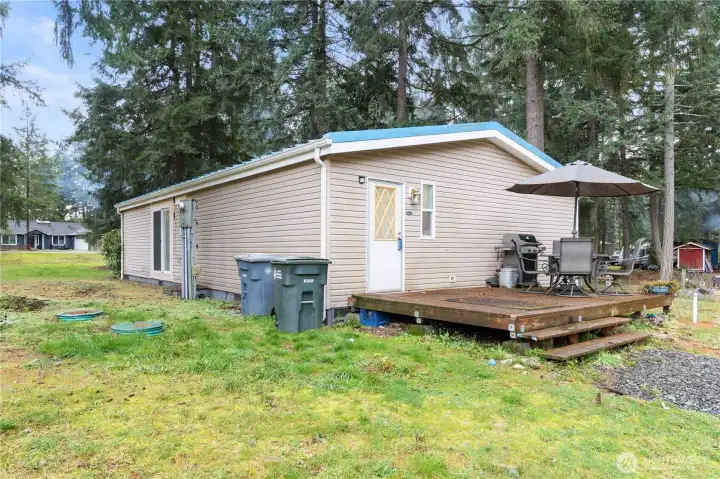 A side view of the home showing the back deck and the open yard surrounding the property. This angle highlights the level outdoor space, the forested backdrop, and the quiet setting. The deck offers plenty of room for outdoor dining and everyday use in a peaceful, natural environment.