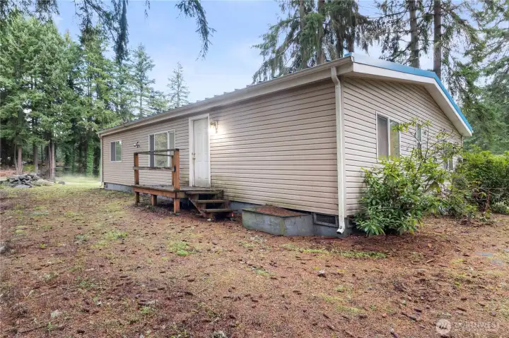 A closer look at the side entry of the home, framed by tall evergreens for added privacy. The natural ground cover and mature landscaping emphasize the wooded setting, while the simple porch provides easy access and a welcoming secondary entrance.