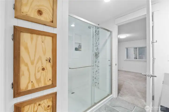 The master bath features a fully tiled walk-in shower with a glass door, accent tile column, and a built-in niche for essentials. Clean white finishes keep the room bright, and the layout offers a straightforward flow into the adjoining bedroom.