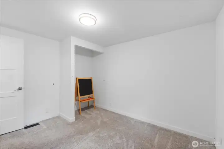 This secondary bedroom offers fresh carpet, a wide window framing the wooded surroundings, and flexible wall space ready for furniture or shelving. The soft neutral palette keeps the room open and inviting, ideal for a guest room, home office, or play space.