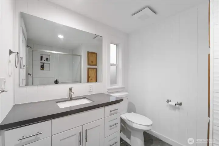 A clean and modern primary bath featuring a wide quartz-topped vanity, full-width mirror, and updated fixtures. The large walk-in shower with sliding glass doors and decorative tile accents adds a polished feel. Bright lighting and simple white paneling keep the space fresh and calming.