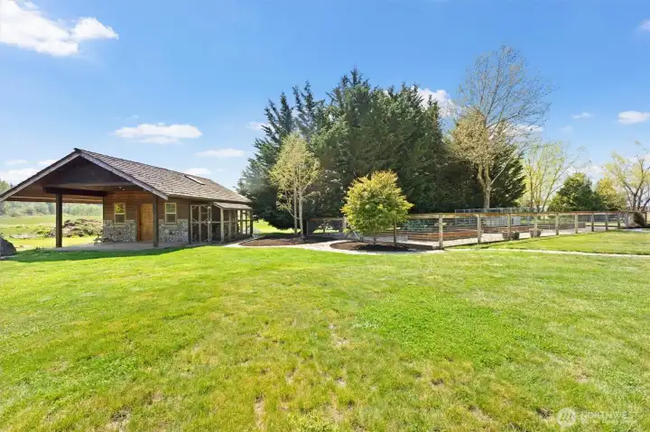 The studio with covered wood storage sits next to a fenced garden area with raised beds plumbed for sprinklers and a dog kennel.