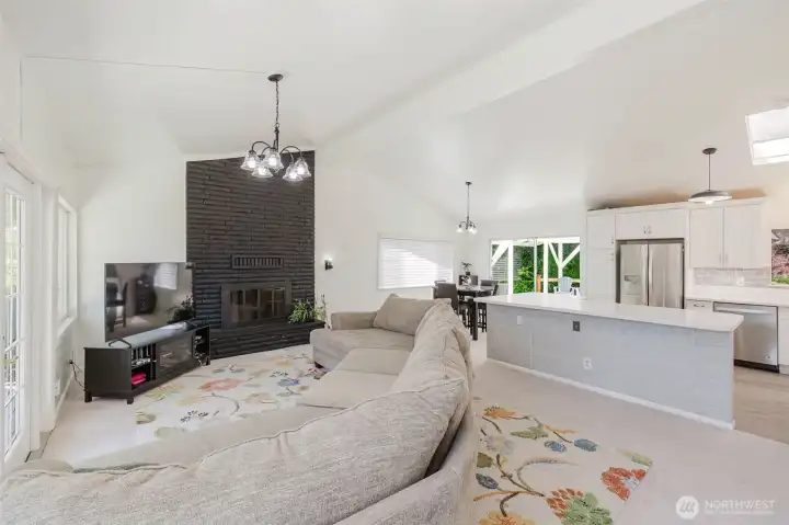 Bright and inviting open-concept living space with vaulted ceilings, abundant natural light, and a striking black brick fireplace that creates a beautiful focal point. Spacious kitchen with oversized island flows seamlessly into the living and dining areas — perfect for entertaining or everyday living. Bright and inviting open-concept living space with vaulted ceilings, abundant natural light, and a striking black brick fireplace that creates a beautiful focal point. Spacious kitchen with oversized island flows seamlessly into the living and dining areas — perfect for entertaining or everyday living.