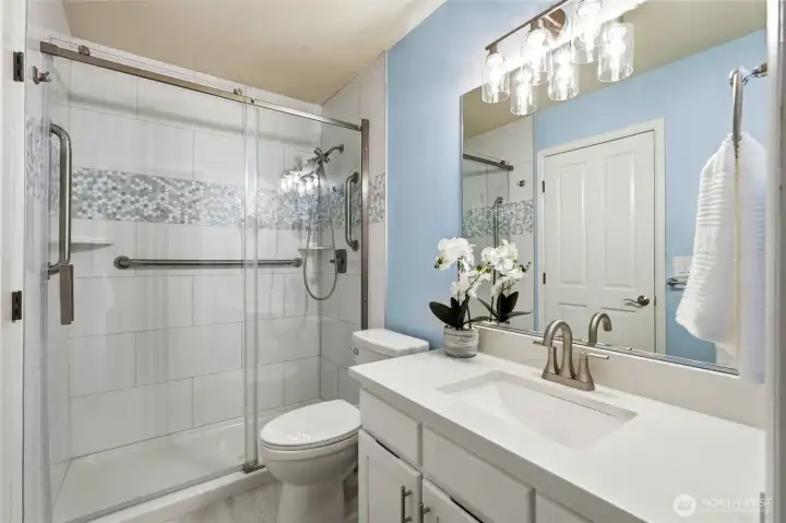 Updated main-level primary bath features a sleek step-in shower with custom tile detail, quartz countertop, modern lighting, and a spacious walk-in closet for added comfort and convenience.