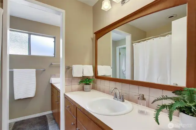 This large bathroom has two sections. The main section has the bath tub/shower and larger vanity/counter space. The farther section is just the toilet and sink, with a smaller vanity, counter and mirror. Beyond the toilet you can see (in the mirror) the door to the primary bedroom.  It's called a "Jack n Jill" bath, when it can be accessed from both bedroom and hallway.