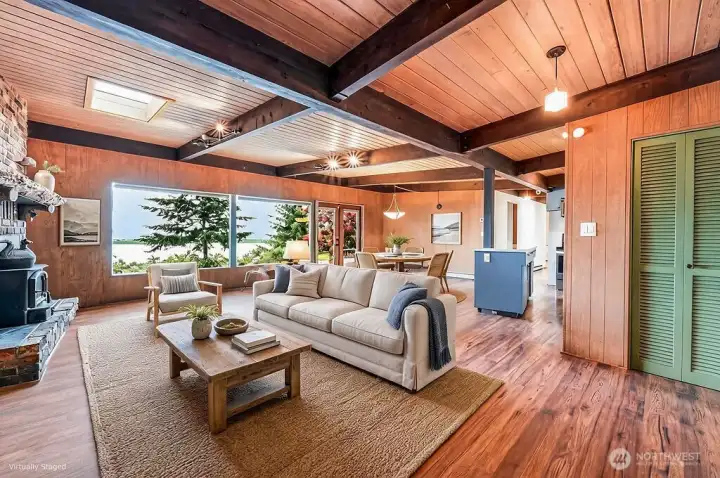 This staged photo of the main living space, with wood beams and ceiling, skylight, and wood burning fireplace. The front door is to the right.
