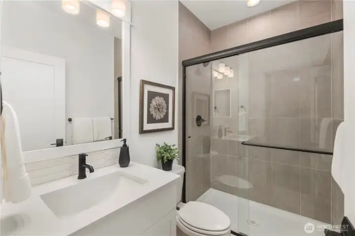 Main-Level Guest Bathroom:  This well-appointed bath features a wide quartz-topped vanity, soft neutral tilework, and a frameless glass shower enclosure accented with matte black hardware. Its thoughtful layout supports comfort and convenience for guests or main-level living. With Gooden Hollow’s Bothell location in the Canyon Park corridor, this space benefits from close proximity to parks, everyday amenities, and major commuter routes. Clean lines and modern fixtures bring a polished, cohesive feel to this main-floor bath.    These photos are from Lot 5 at Gooden Hollow, the community model. The home listed will have changes, so all photos, renderings, site maps, and floorplans are for visualization purposes only. Plans, colors, building orientation, finishes, home outlook, and other details can and WILL vary.