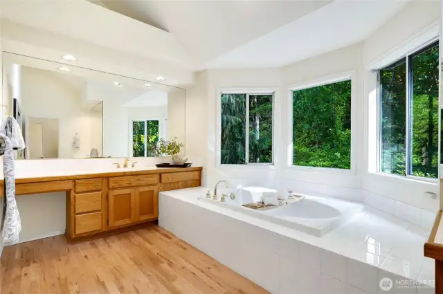 Unwind in the spa-like primary bath featuring a deep soaking tub surrounded by picture windows that frame peaceful, wooded views.