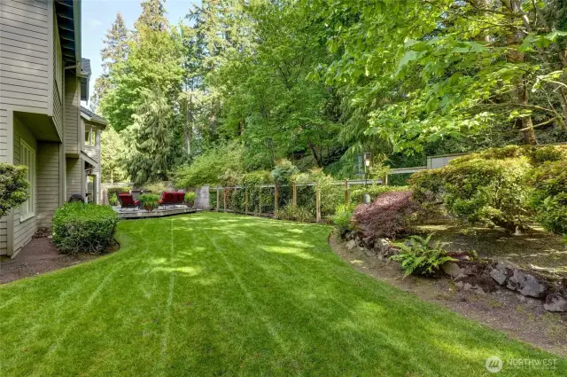 Large fully fenced backyard with a beautifully manicured lawn, mature landscaping, and forested backdrop. Ideal for outdoor play, pets, or peaceful relaxation.