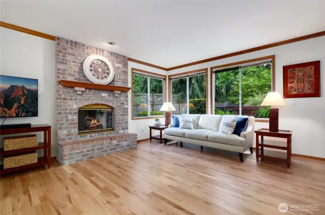 Charming family room with a classic brick fireplace and large picture windows that frame lush greenery. Gleaming hardwood floors and natural light create a warm, inviting space—perfect for relaxing.