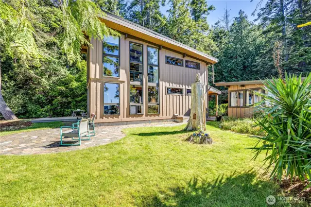 Side of the home with small patio, grassy area and out building beyond. Beautiful light here.