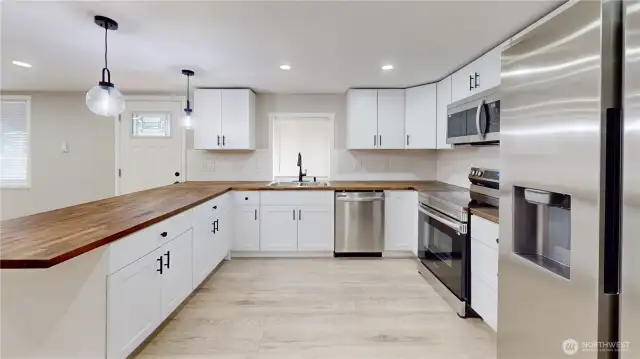 Bright and stylish kitchen featuring upgraded countertops, sleek tile backsplash, and stainless-steel appliances—including a flattop range. White easy-close cabinets and drawers offer both elegance and convenience, while modern light fixtures add a fresh touch. A spacious breakfast bar provides the perfect spot for casual dining or entertaining.