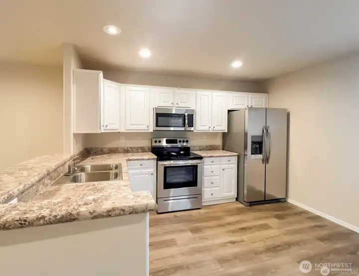 The kitchen is open with room for dining space and featuring all stainless steel appliances, white cabinets providing ample storage, sleek counters, and modern wood design vinyl flooring.