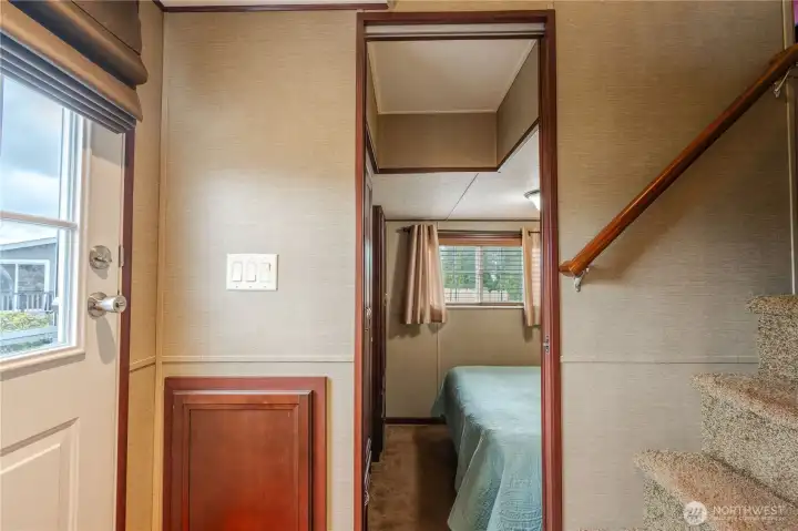 The back door opens to a hallway. To the right is the bathroom, kitchen, dining and living rooms. To the left is the primary bedroom. Straight ahead are the stairs leading to a second bedroom and den.