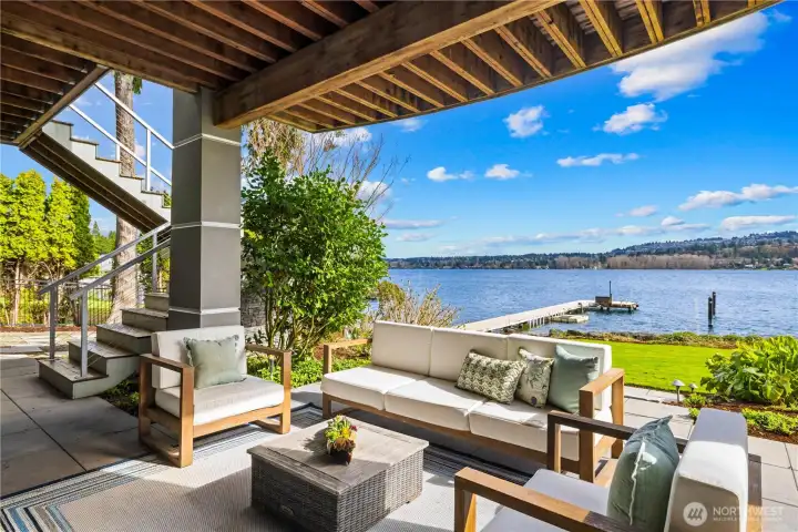 Covered waterfront patio with exposed timber ceiling and comfortable outdoor seating overlooking the private dock, manicured lawn, and sweeping lake views. The perfect spot to relax and take in the scenery year-round.