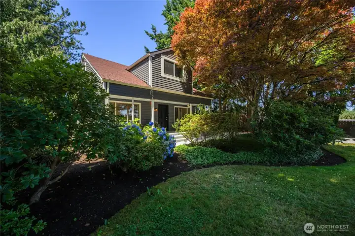 Charming 1920s home nestled on a beautifully landscaped corner lot- framed by mature trees, vibrant hydrangeas, and lush greenery for a peaceful, garden setting.