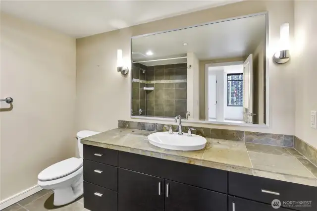 The bathroom features a tiled shower-tub combination with earthy stone finishes, complementing the vanity design