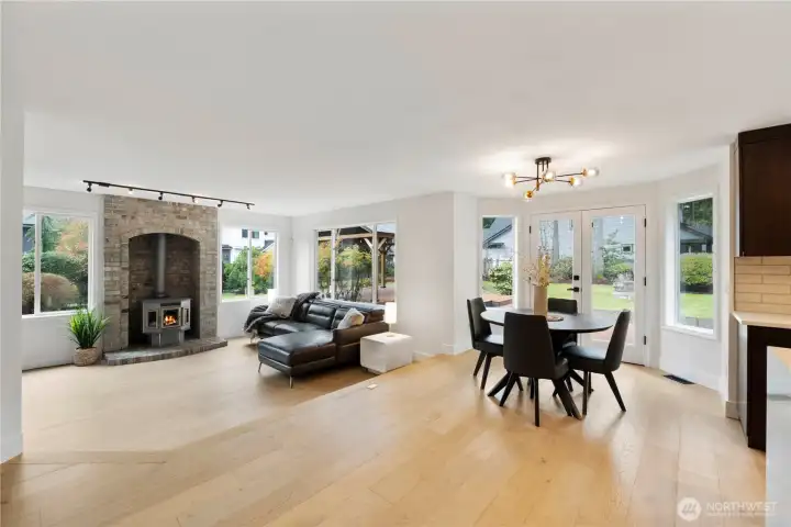 Bright and inviting open-concept living and dining space featuring wide-plank hardwood floors and abundant natural light. The cozy brick-accented pellet fireplace creates a warm focal point, while large windows and glass doors seamlessly connect indoor living to the private backyard. The modern dining area is perfect for entertaining, complemented by stylish lighting and a clean, contemporary design. A perfect blend of comfort, functionality, and indoor-outdoor living.