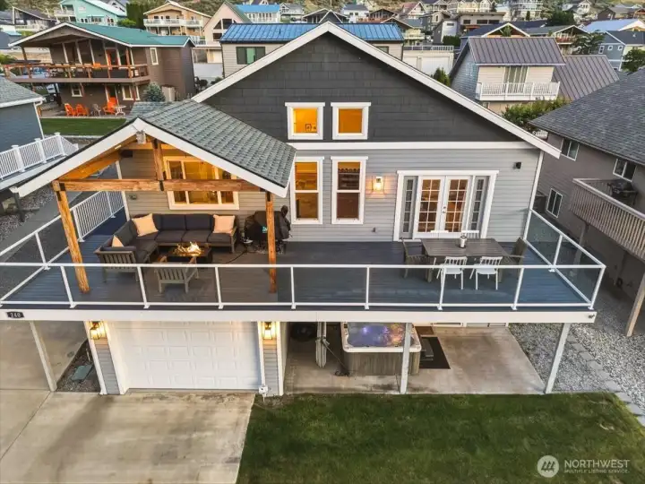 Aerial view of home and river and mountain view deck and outdoor living space with lounge area and fire table, dining and BBQ area and grassy lawn.