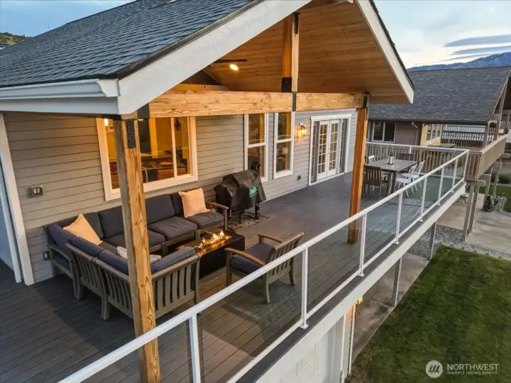 Twilight view of river and mountain view deck and outdoor living area with lounge area with fire table, outdoor dining table and lower level with private hot tub and grassy lawn.