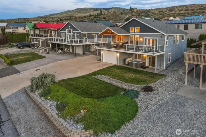 Twilight view of home with large, grassy lot, driveway, river and mountain view deck and attached oversized garage designed to house a wakeboarding boat, golf cart, exercise equipment and more.