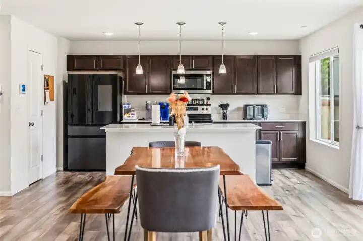 Bright, open-concept kitchen featuring a center island with seating, pendant lighting, black stainless refrigerator, and a sunny dining area.