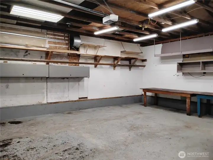 Plenty of lighting in this garage which has a automatic door opener, permanent mounted shelves and several movable workbench tables.