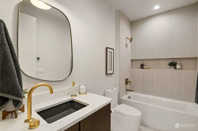 Secondary bathroom with gold fixtures, quartz countertop, textured tile accents, and a tub-shower combo with built-in shelving.