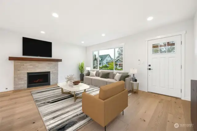 The living area with recessed lighting features a cozy gas fireplace with solid wood mantle, ductless heating with air conditioning, and brand new beautiful wide planked wood floors underfoot.  Notice the large front window allowing a ton of light to pour inside.  There is a smurf tube above and to the right of the fireplace so no tangled cords are hanging down under the TV.