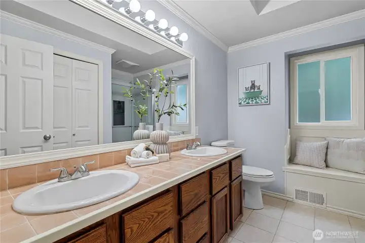 The primary bath features double sinks, tile flooring, a built-in bench seat beneath the window, and a skylight that fills the space with natural light.