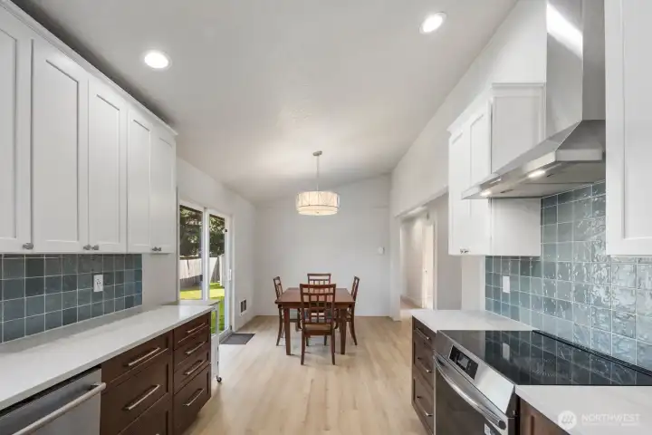 Remodeled kitchen with quartz countertops, induction range, stainless vent hood, two-tone cabinetry, blue tile backsplash, and open view to dining area with sliding glass door.
