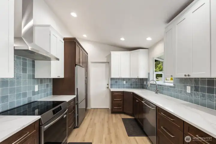 Remodeled kitchen with quartz countertops, induction range, stainless steel vent hood, two-tone cabinetry, blue tile backsplash, and stainless steel appliances.