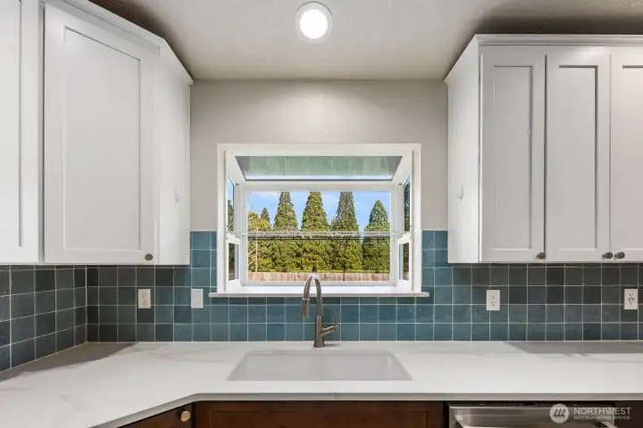 Kitchen with under-mount sink, quartz countertops, full tile backsplash, upper cabinetry, and window overlooking the backyard.