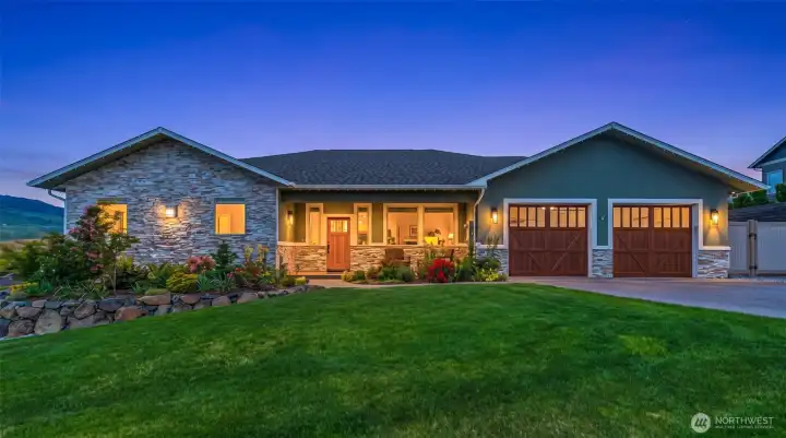 The exterior showcases rich stone accents, warm architectural lighting, and carriage-style wood garage doors.
