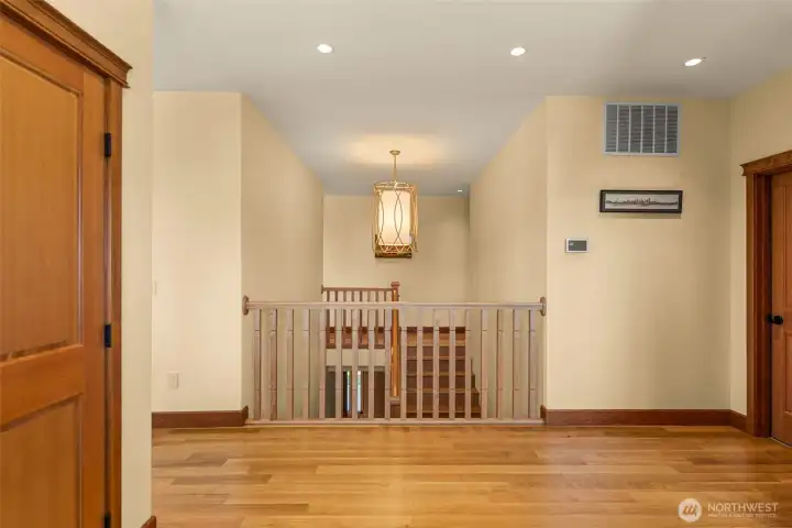 Clean architectural lines and warm natural finishes define this upper-level landing, featuring rich hardwood flooring, solid wood trim and doors, recessed lighting, and custom railing details. The open stairwell design creates an airy sense of space while the statement pendant light adds warmth and visual balance.