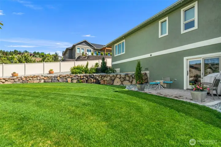 Beautifully maintained backyard designed for both relaxation and low-maintenance enjoyment, featuring expansive lush lawn, custom rock retaining walls, and thoughtfully curated landscaping