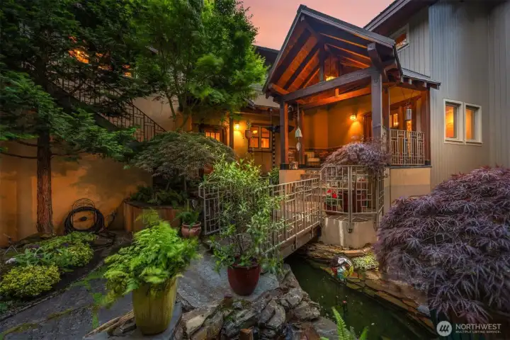 Approach front door over bridge with multilevel fountain and lush greenery.
