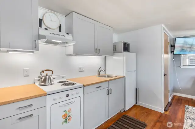 Compact kitchen with white cabinets, laminate butcher block look counters, open shelving, and full-size fridge.