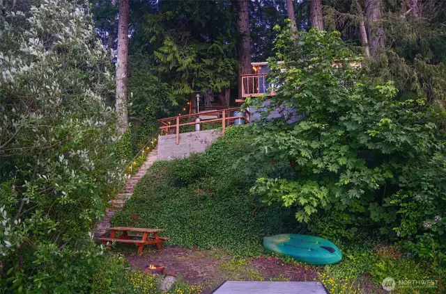Lakeside landing area at dusk with soft lighting around the firepit, picnic table, and kayak storage.