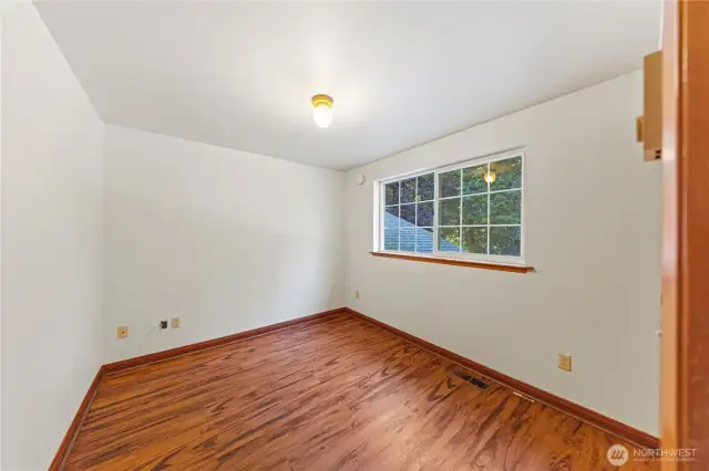 Bright guest bedroom with wood-style flooring and a large window overlooking the treed property.
