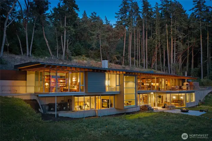 As evening falls, the residence glows warmly against the forested backdrop, highlighting the refined architecture and expansive glass walls that define this exceptional San Juan Island luxury waterfront estate.