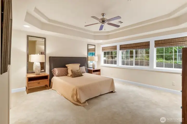 Spacious primary suite featuring tray ceilings, a ceiling fan, and a wall of windows overlooking the peaceful forest setting, creating a bright and relaxing retreat.