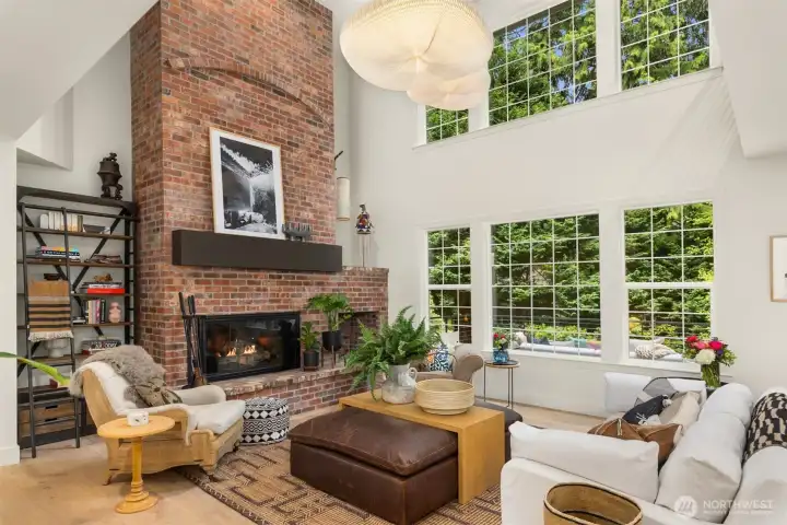 A classic brick fireplace grounds the living room, while picture-grid windows frame lush Pacific Northwest greenery like living artwork beyond the glass.
