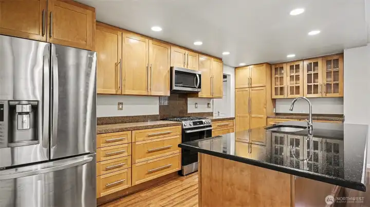 Modern kitchen with natural toned cabinetry. Spacious island features a sink, dishwasher, outlet, and plenty of storage—perfect for meal prep and casual gatherings.