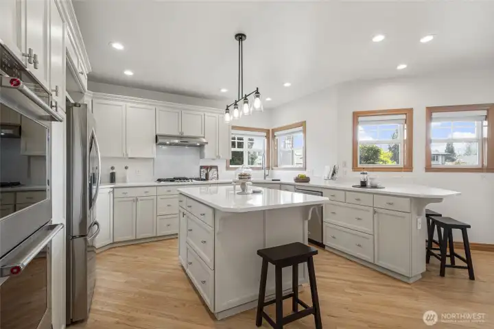 Spacious kitchen featuring quartz countertops, expansive island, stainless steel appliances, abundant cabinetry, and warm wood-wrapped windows filled with natural light.