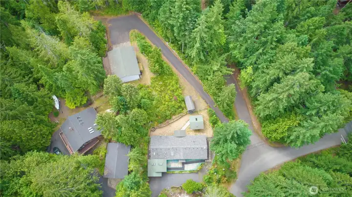 An aerial birds-eye view highlighting the layout of the property, showing the main home, the detached garage/shop, outbuildings, and the private, paved driveways surrounded by a lush canopy of mature trees.