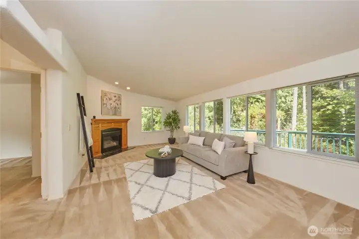 The bright and airy living room features a cozy wood-framed fireplace, plush neutral carpeting, and a wall of large windows that frame peaceful views of the surrounding forest and deck.