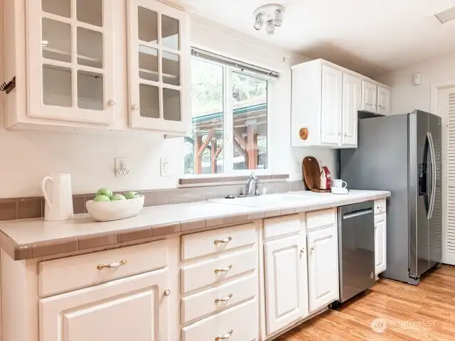 Prepare meals with a view! The kitchen features classic tile countertops, crisp white cabinetry, stainless steel appliances, and a window framing the tranquil, wooded backyard.