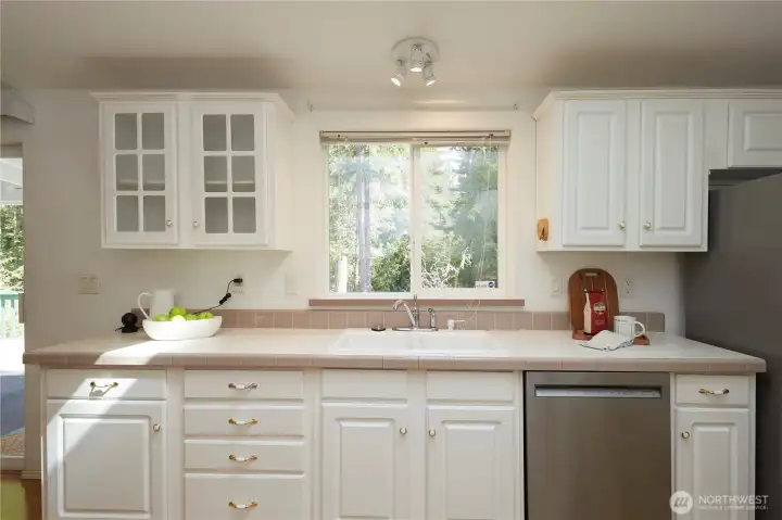 A bright and clean kitchen workspace featuring classic tile countertops, crisp white cabinetry with elegant glass-front accents, stainless steel appliances, and a window overlooking the peaceful forest setting.
