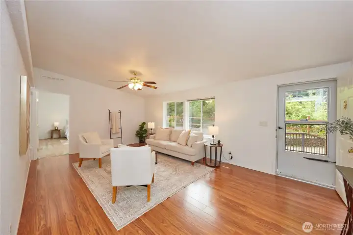The expansive and airy living room acts as the heart of the home, boasting stunning hardwood-look flooring, a classic ceiling fan, and large windows that seamlessly blend indoor comfort with the beautiful surrounding nature.